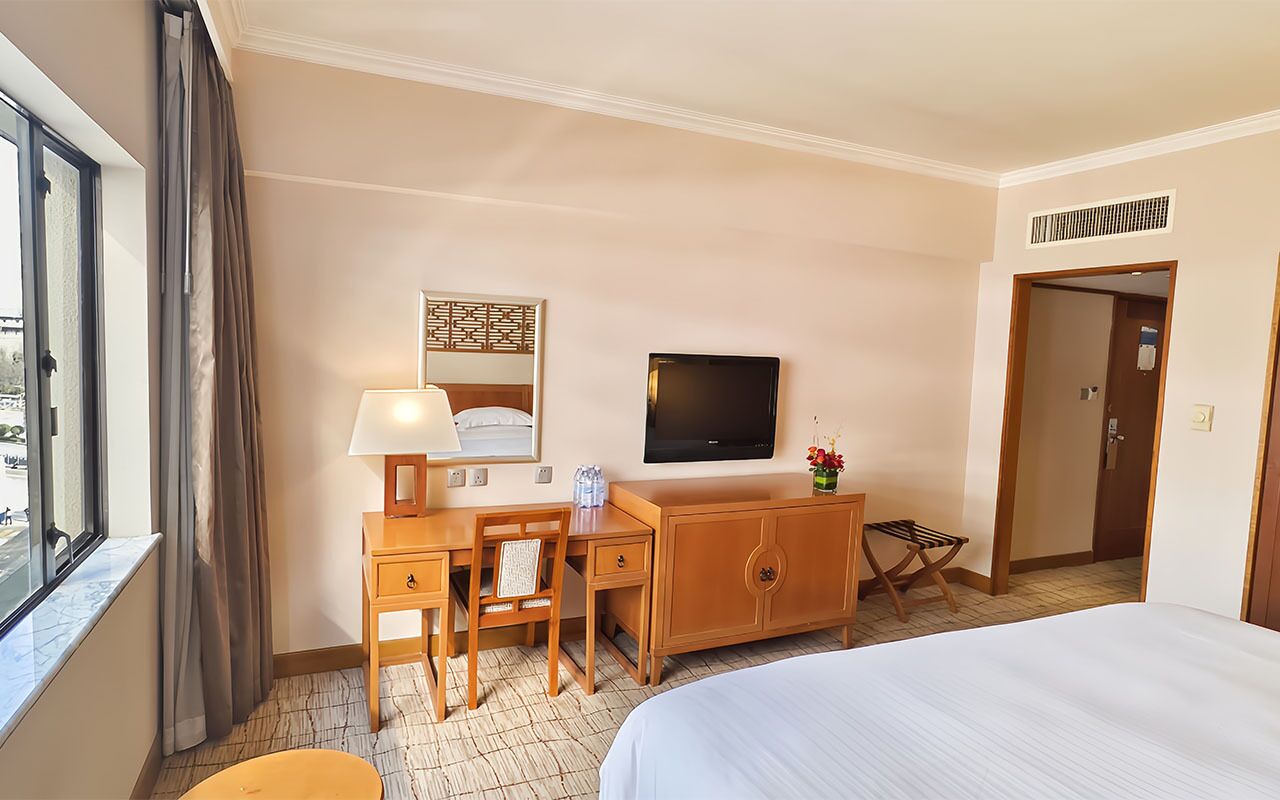 A hotel room with a bed, a desk with a chair, a television, a small table with a lamp, and a window with curtains.