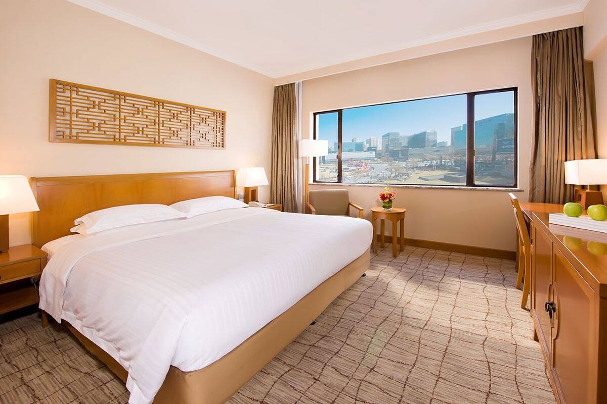 Hotelkamer met een groot bed, een bureau, een stoel, uitzicht op de stad en een decoratief kunstwerk aan de muur.