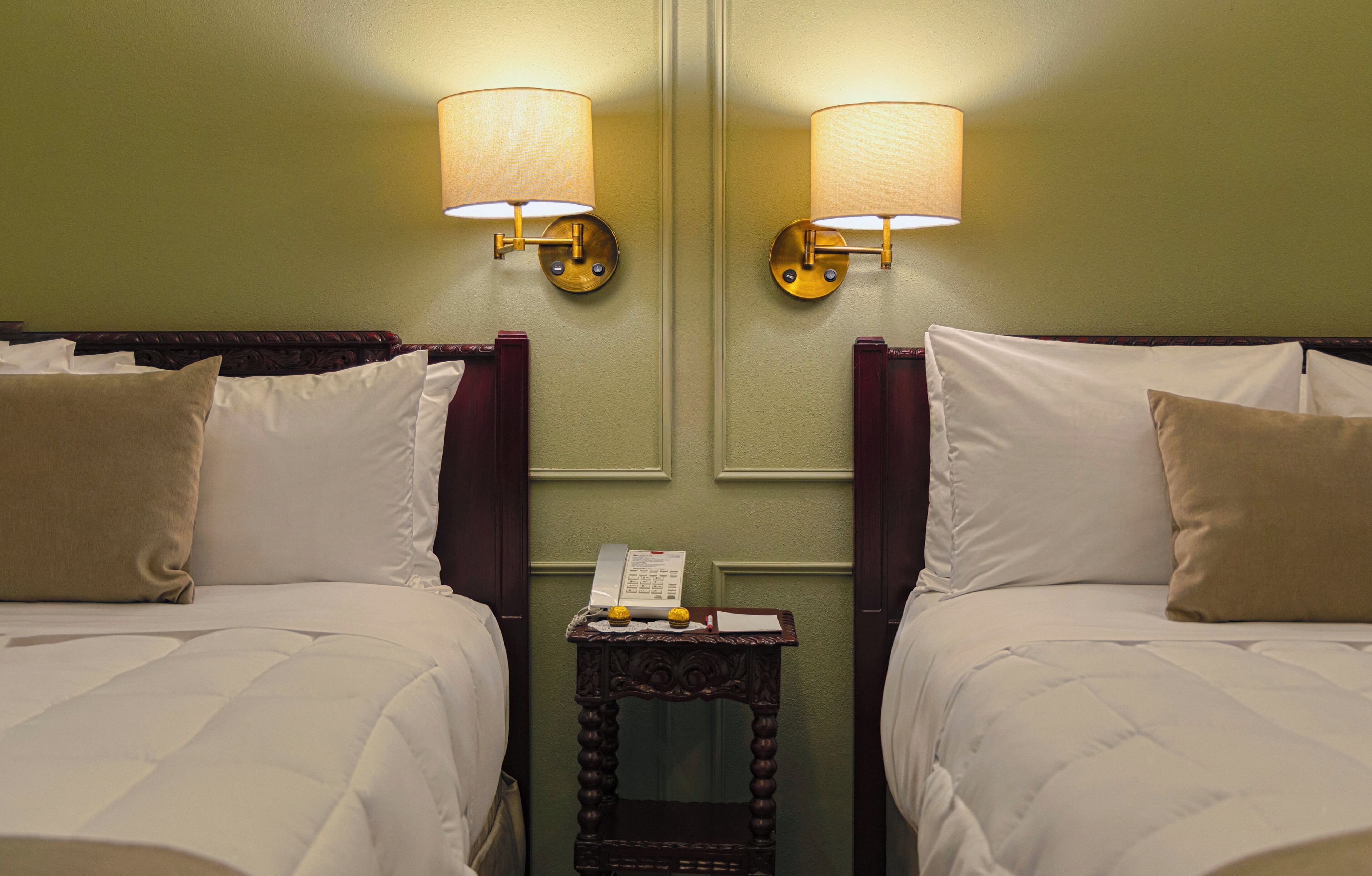 A hotel room with two beds, a nightstand with a telephone, and wall-mounted lamps.