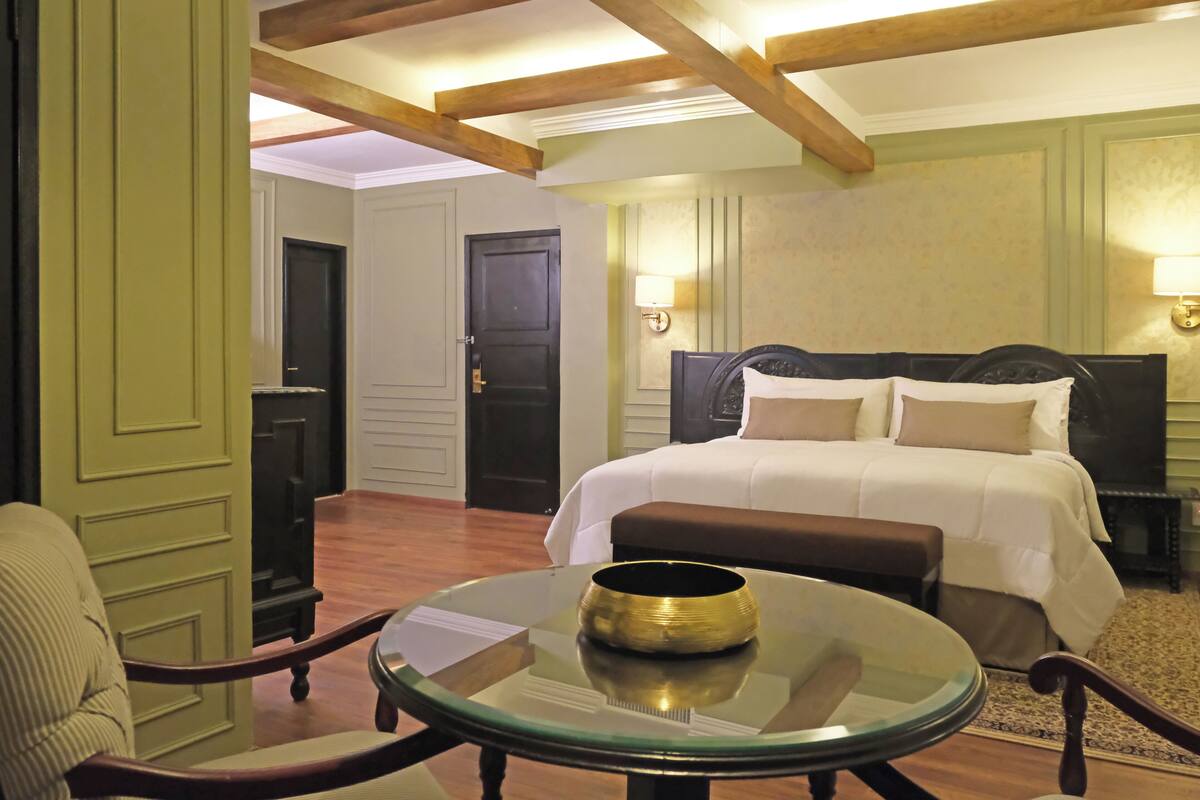 Une chambre d’hôtel avec un lit, une table ronde en verre sur laquelle se trouve un bol, une chaise et une porte au fond.