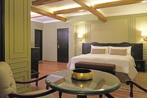 A hotel room with a bed, a round glass table with a bowl, a chair, and a door in the background.
