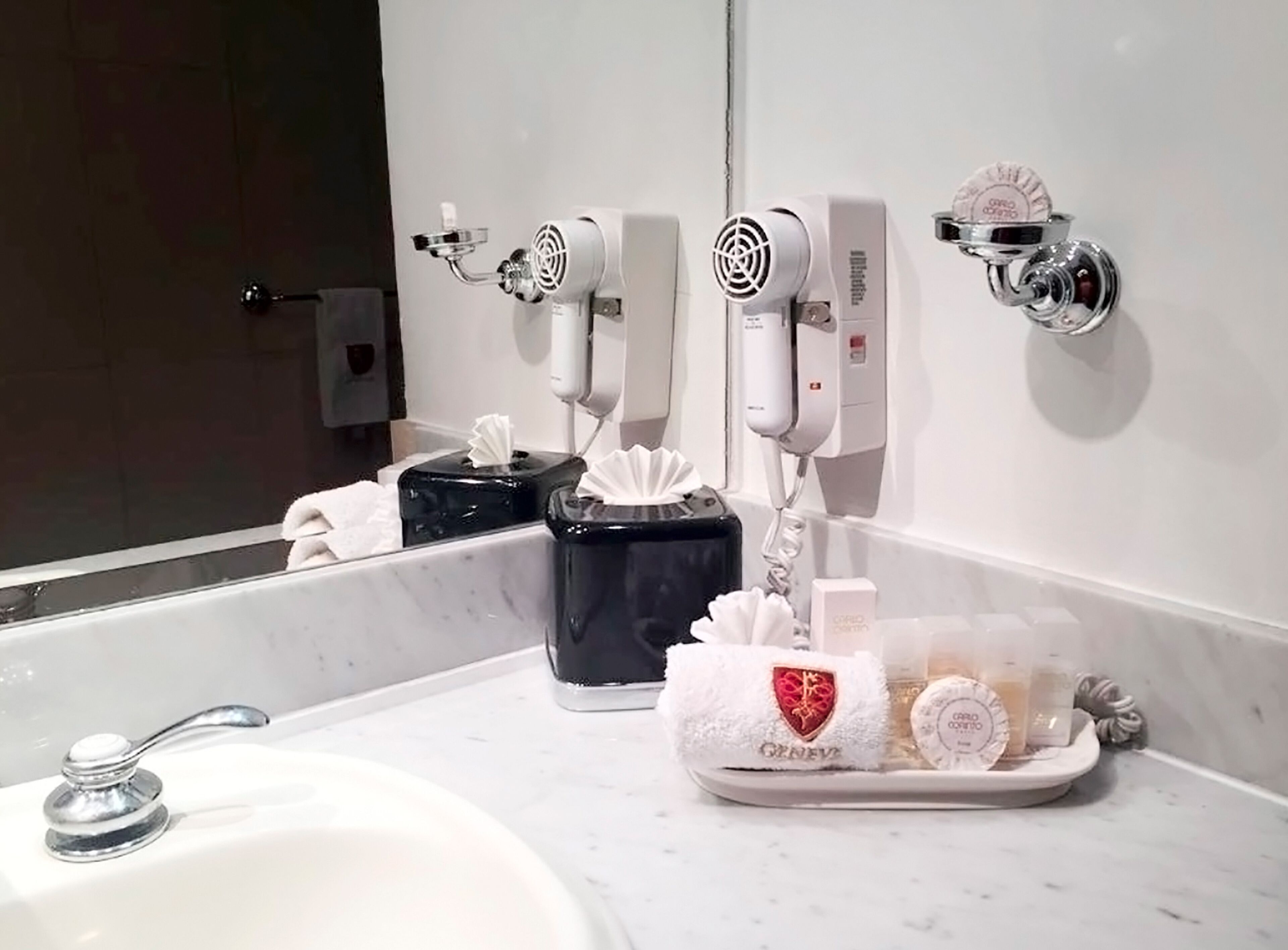 Un baño con un lavabo blanco, un espejo y tres secadores de pelo fijados a la pared.