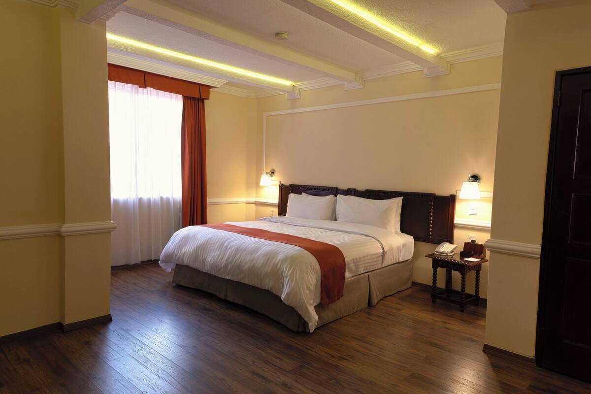Une chambre d’hôtel avec un grand lit, une table de chevet, une lampe et une fenêtre avec des rideaux.