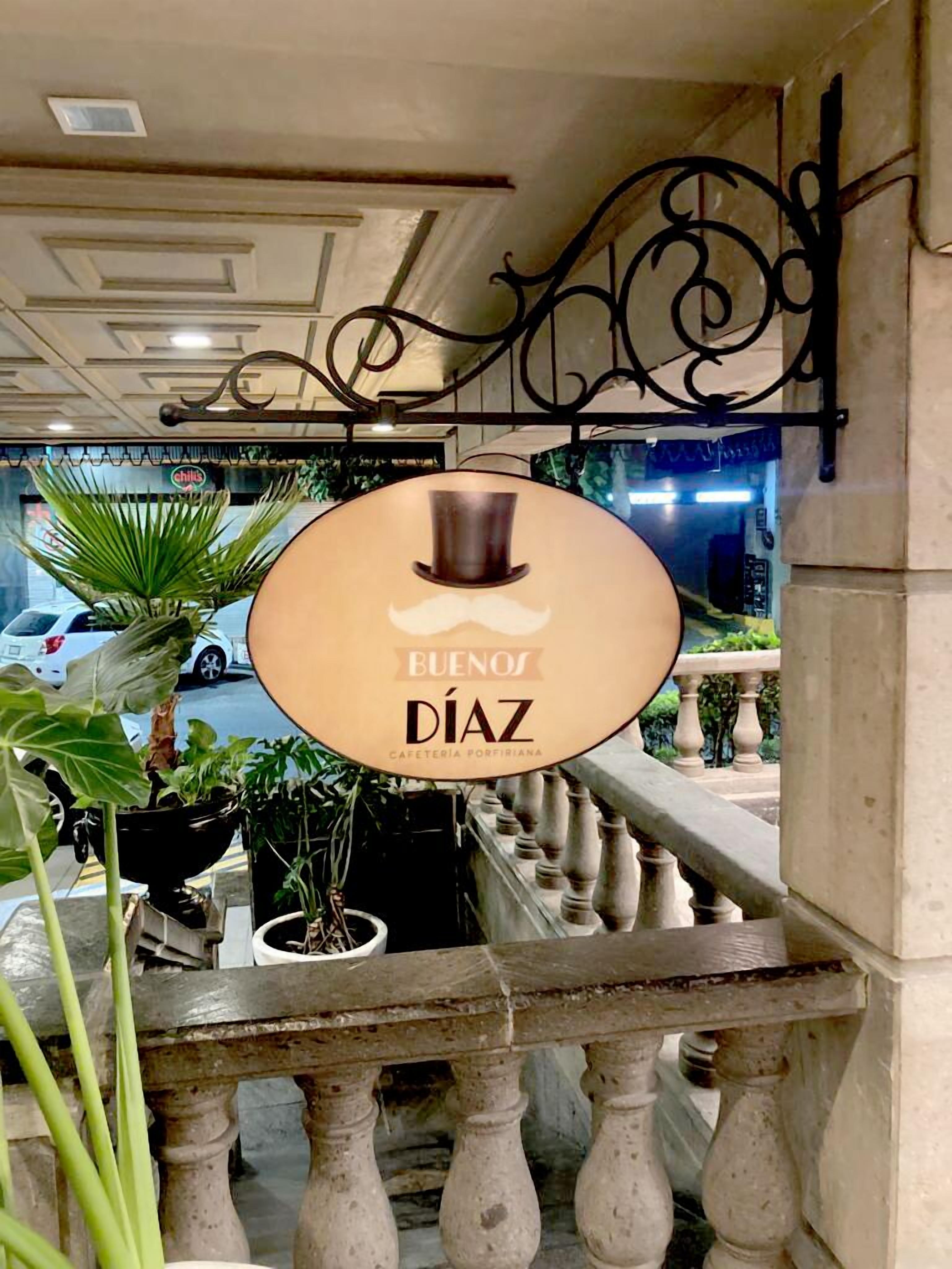 A sign for 'Buenos Díaz' café, with a mustache graphic, located in a building with a balcony and potted plants.