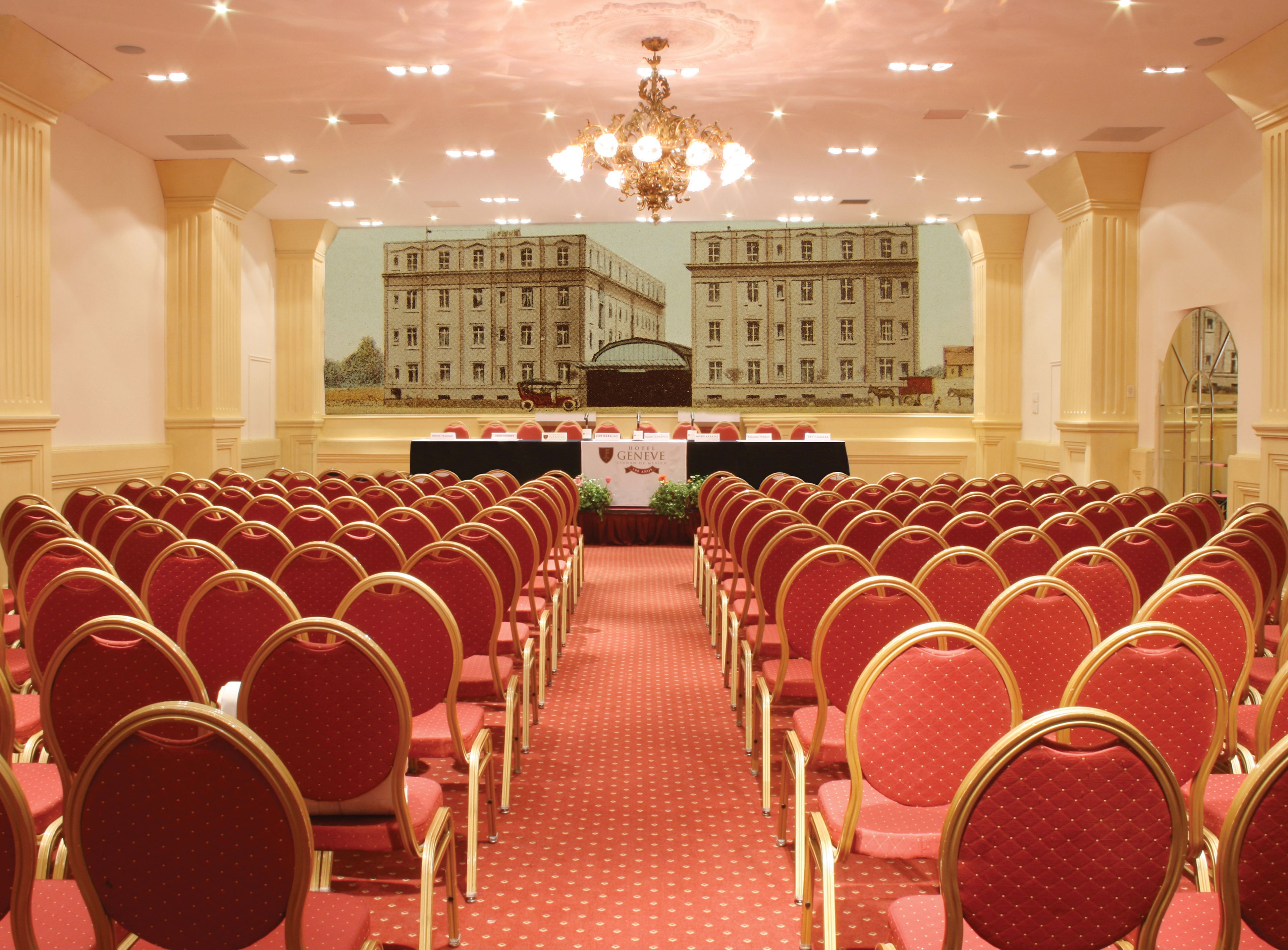 Un salón de conferencias con hileras de sillas rojas, un escenario con un cartel y una araña.