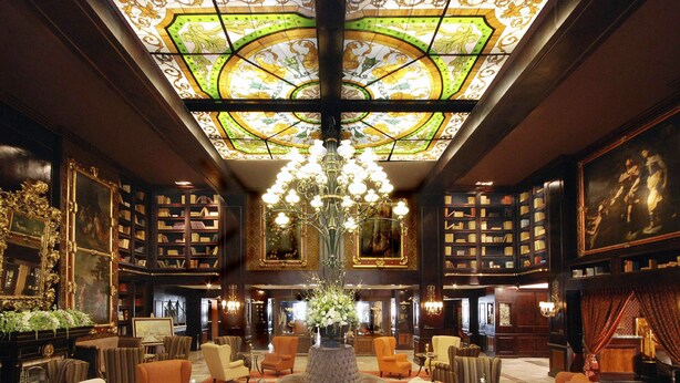 A grand lobby with a large chandelier, ornate ceiling, and a central round table surrounded by sofas and armchairs.