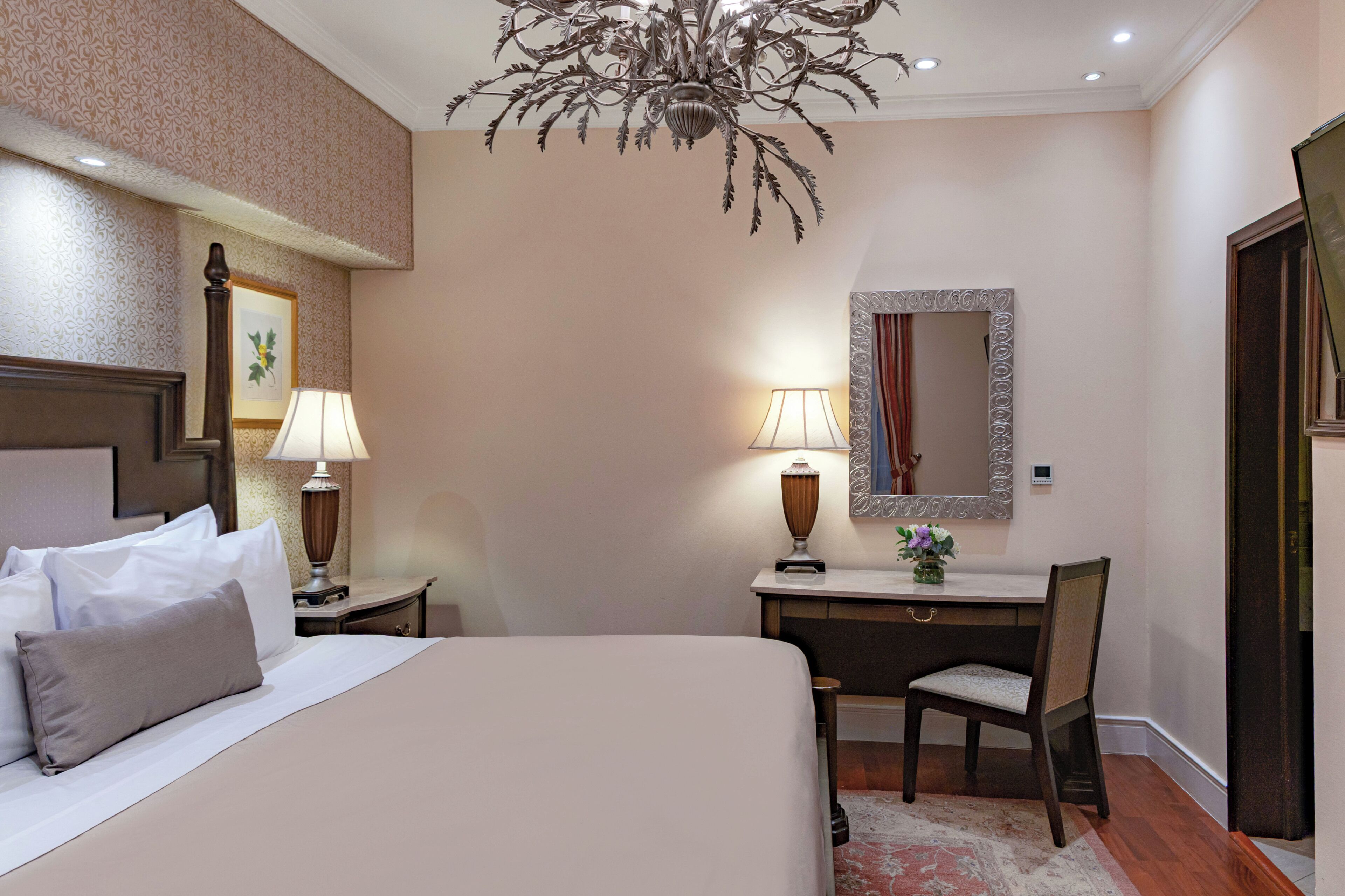 A hotel room with a bed, two bedside tables with lamps, a desk with a chair, a mirror, and a chandelier.