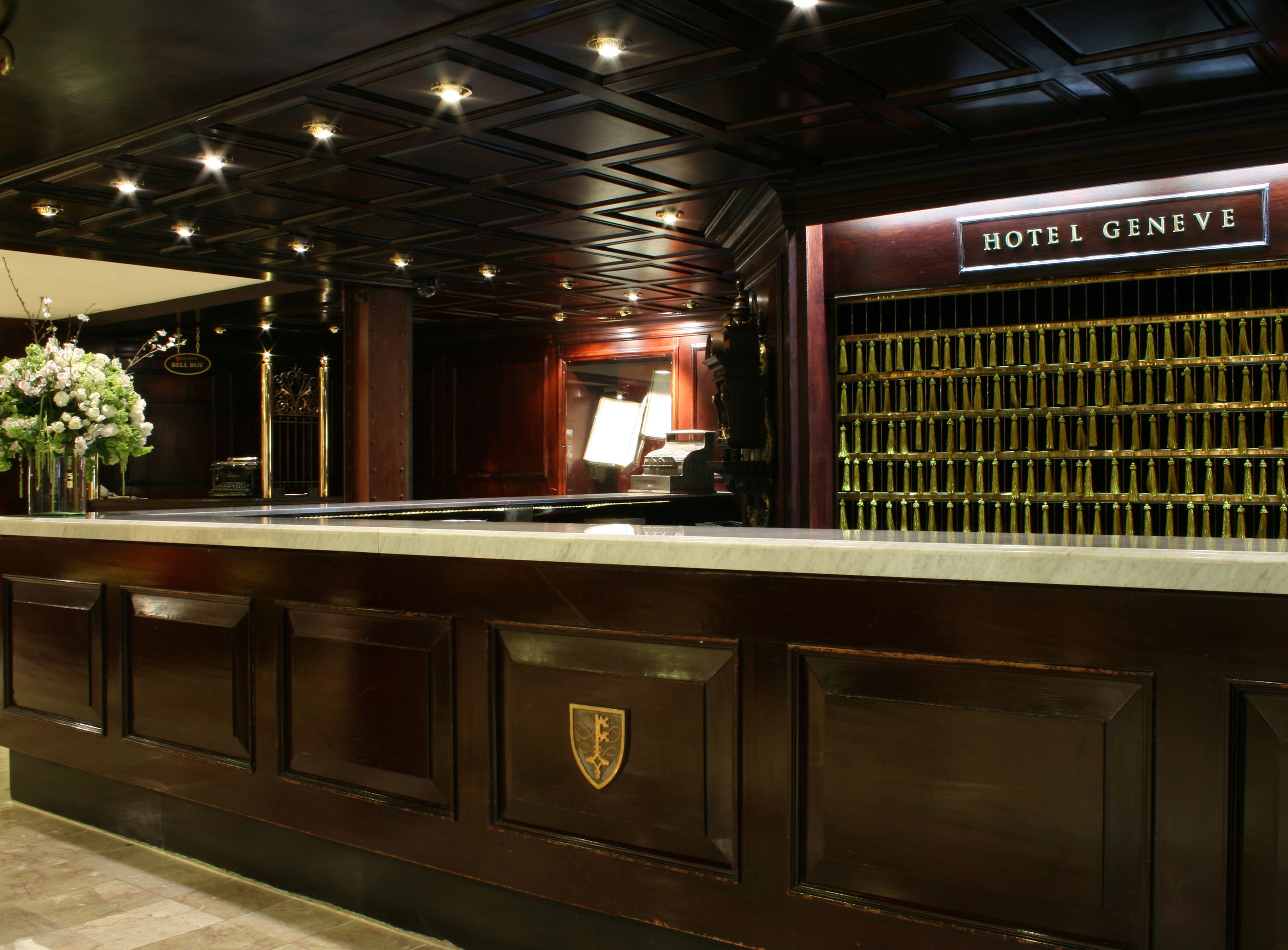 Un'area reception con un bancone in marmo, un cartello con la scritta 'HOTEL GENEVE' e una parete decorativa a griglia.
