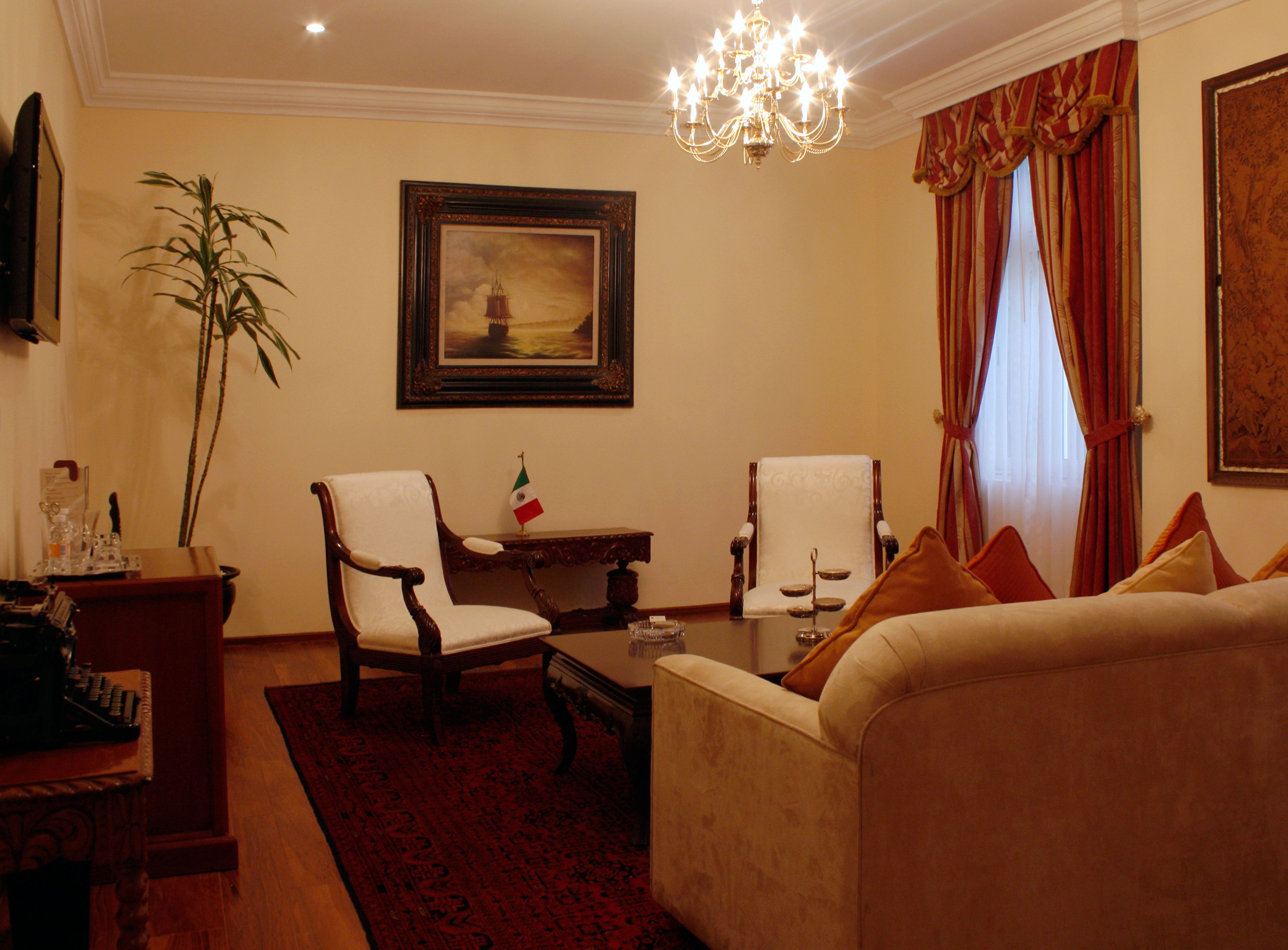 A living room with a sofa, two armchairs, a coffee table, a chandelier, a painting, a flag, and a television.