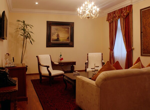 A living room with a sofa, two armchairs, a coffee table, a chandelier, a painting, a flag, and a television.