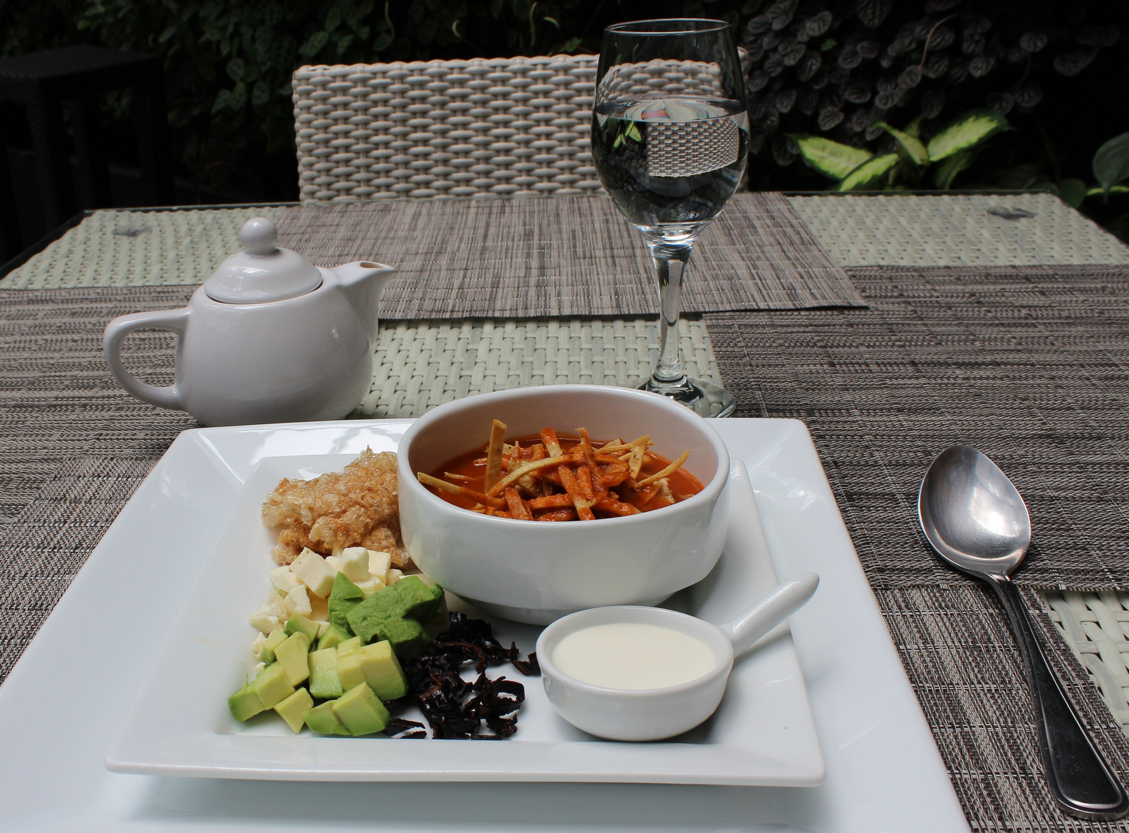 A dining table with a white plate of food, a bowl of soup, a glass of water, a teapot, and a spoon.