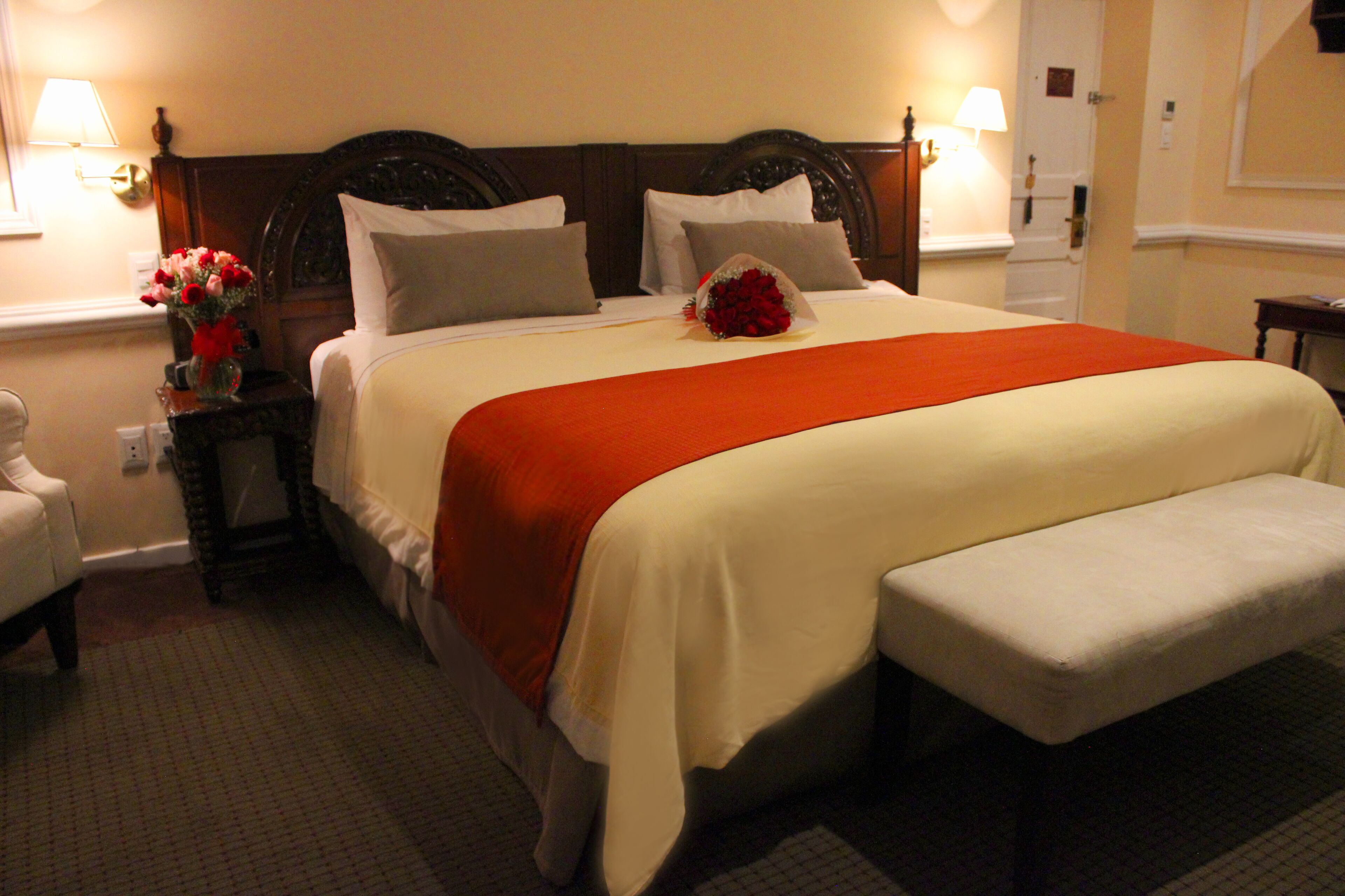 A neatly made bed with a red blanket, a bedside table with a vase of flowers, and a bench at the foot of the bed.