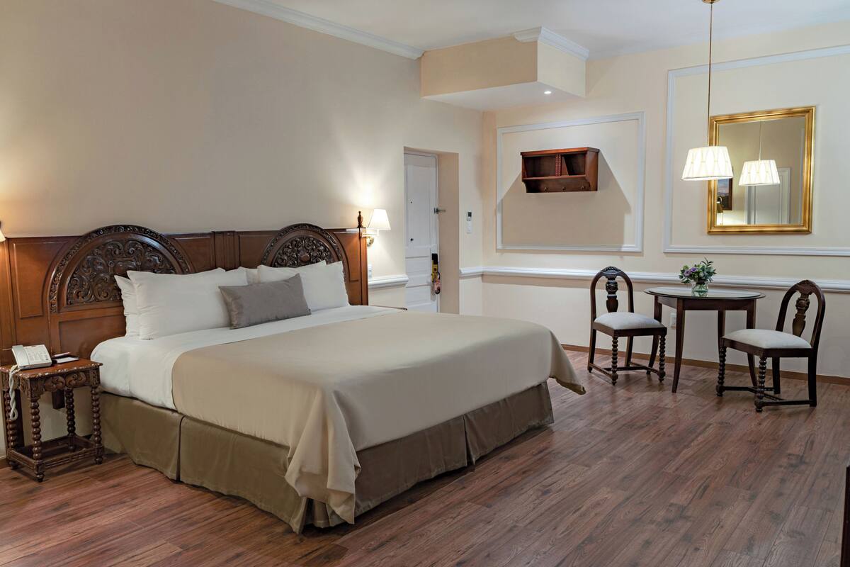 Une chambre d’hôtel avec un grand lit, une table de chevet avec une lampe, une petite table avec des chaises et un miroir au mur.