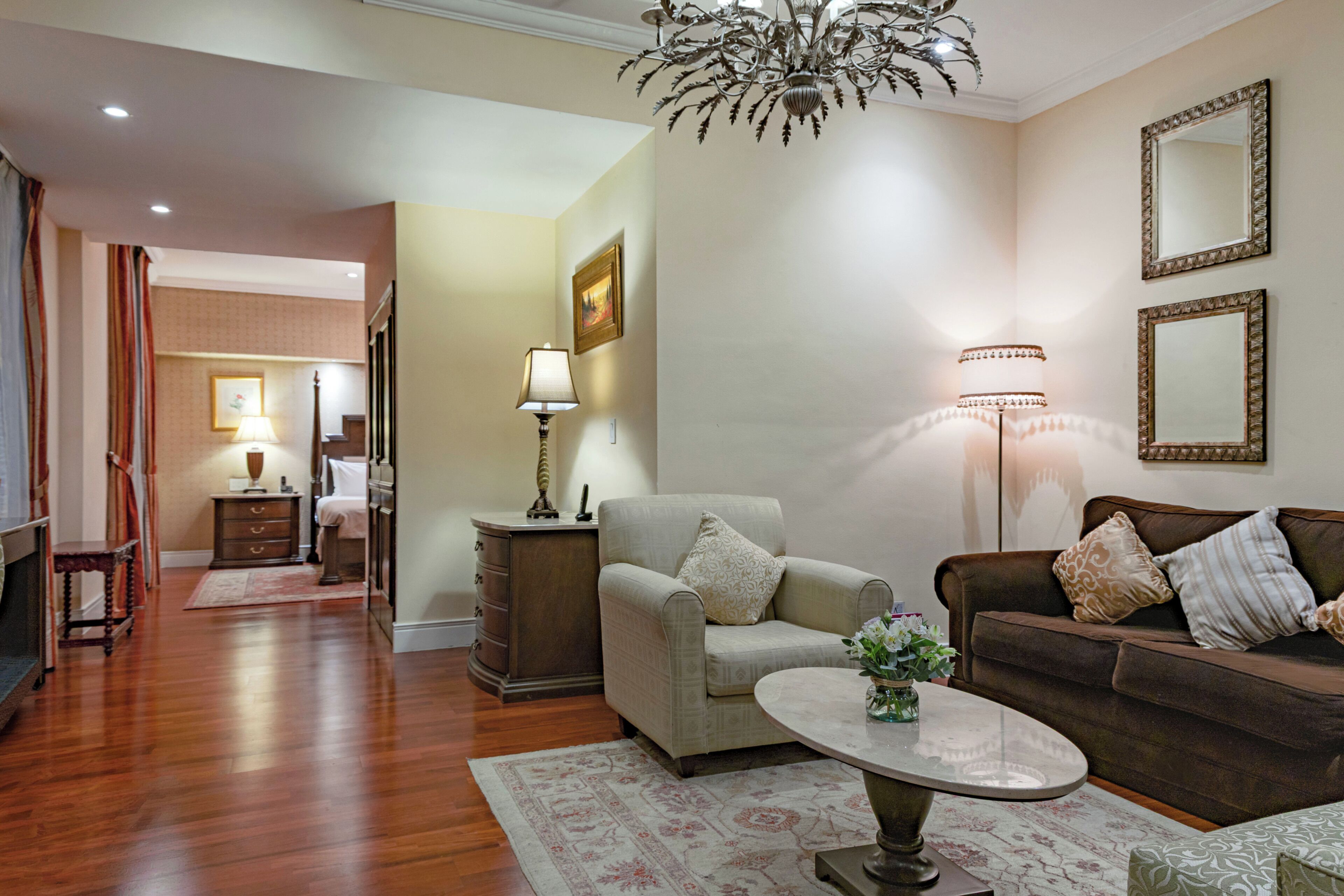 Un salon comprenant un canapé, un fauteuil, une table basse et des lampes. On y trouve également un lustre, un miroir et un tableau accroché au mur.