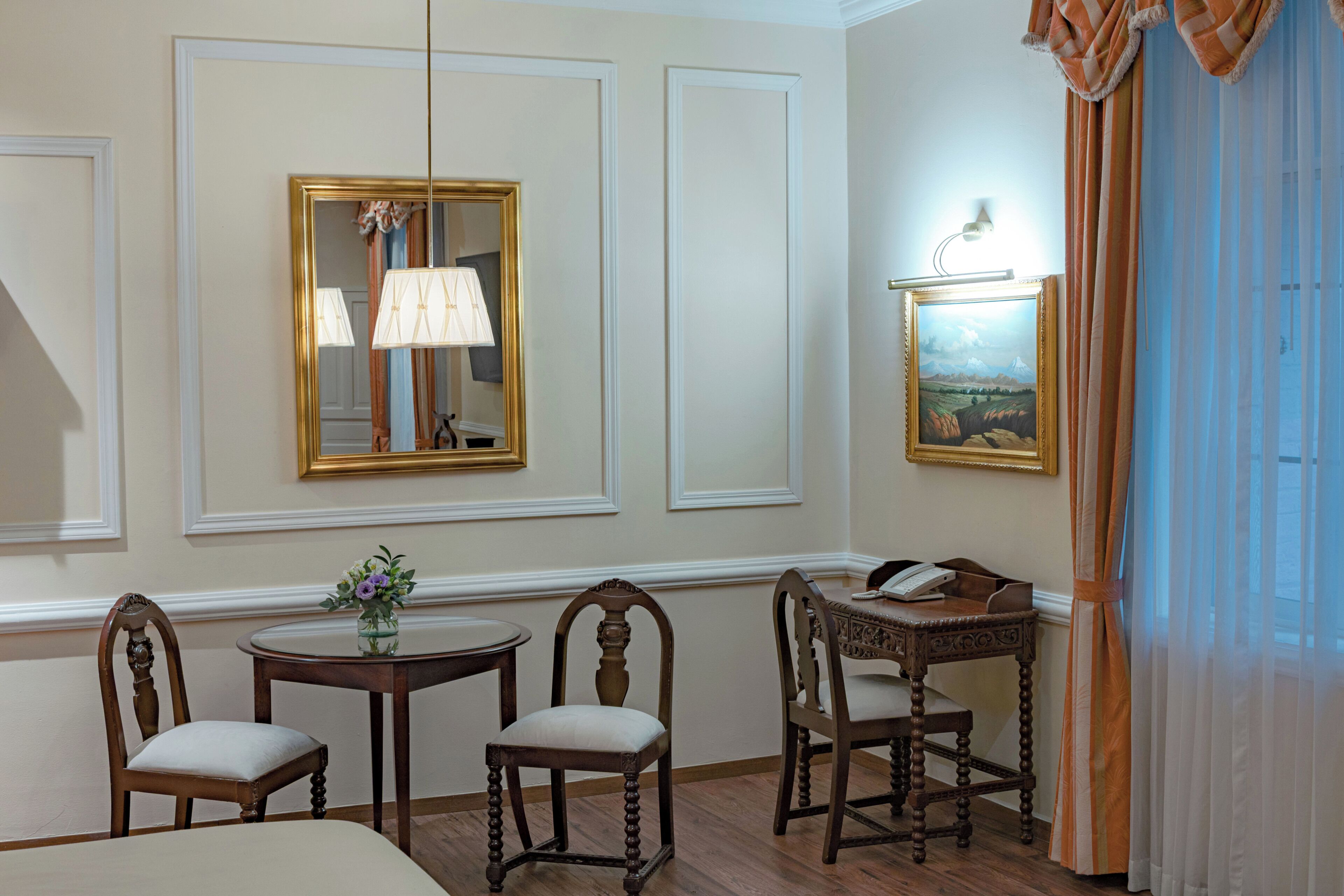 A room with a dining table, two chairs, a lamp, a painting, and a telephone on a desk.