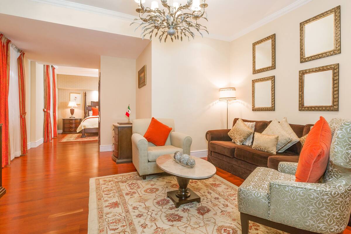 Un salon spacieux avec un canapé marron, un fauteuil gris et un tapis à motifs. On y trouve un lustre, une lampe et un drapeau posés sur une table d’appoint.