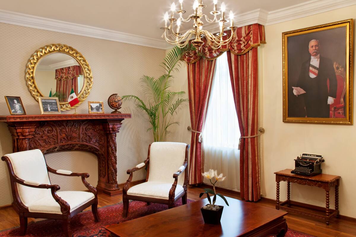 Une pièce élégante, avec deux fauteuils richement décorés, une table en bois, un bureau orné d’un globe terrestre et de tableaux encadrés, un grand miroir, un lustre et un portrait accroché au mur.