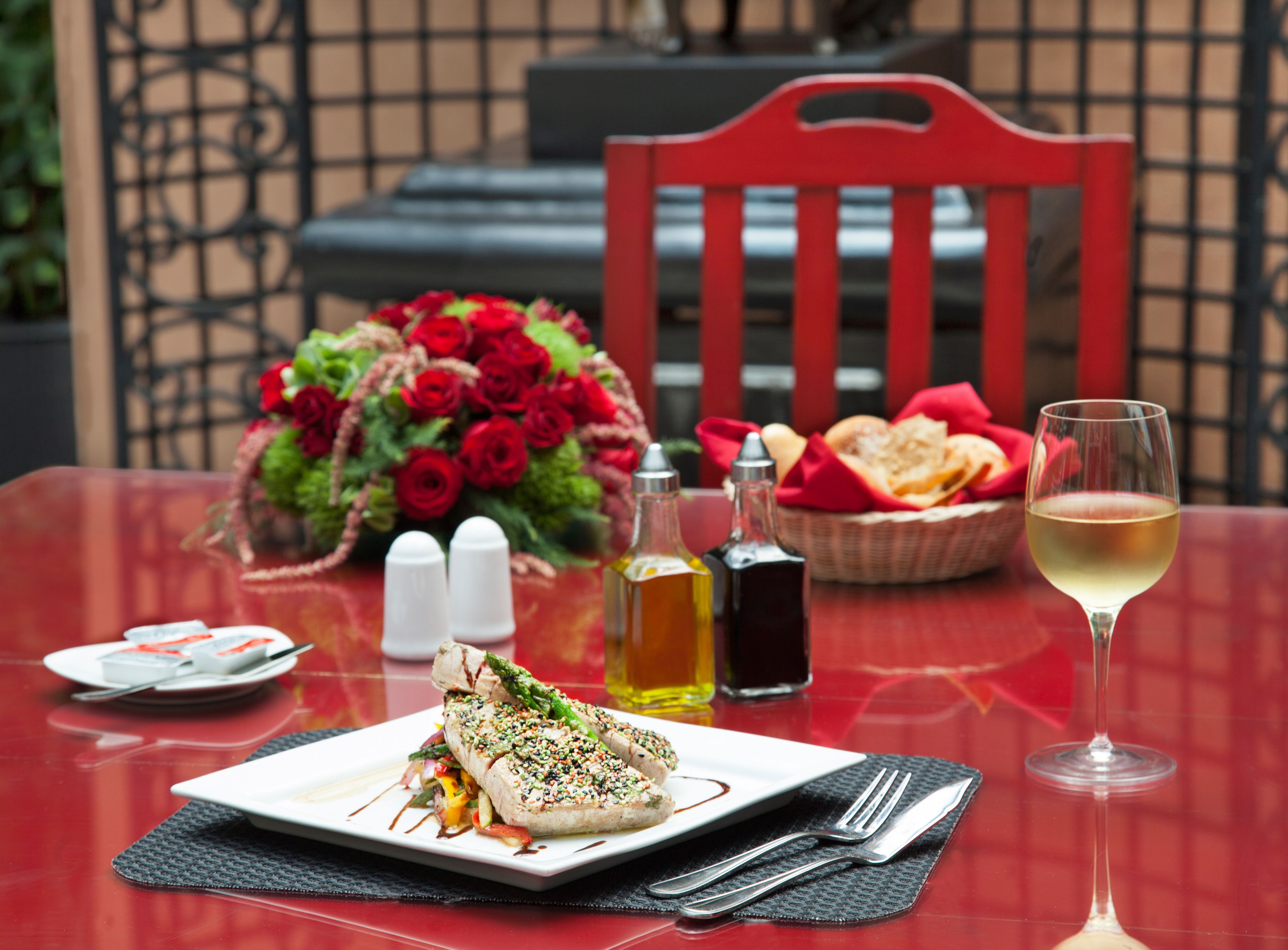 A dining table with a plate of food, a glass of wine, salt and pepper shakers, a basket of bread, and a bouquet of red roses.