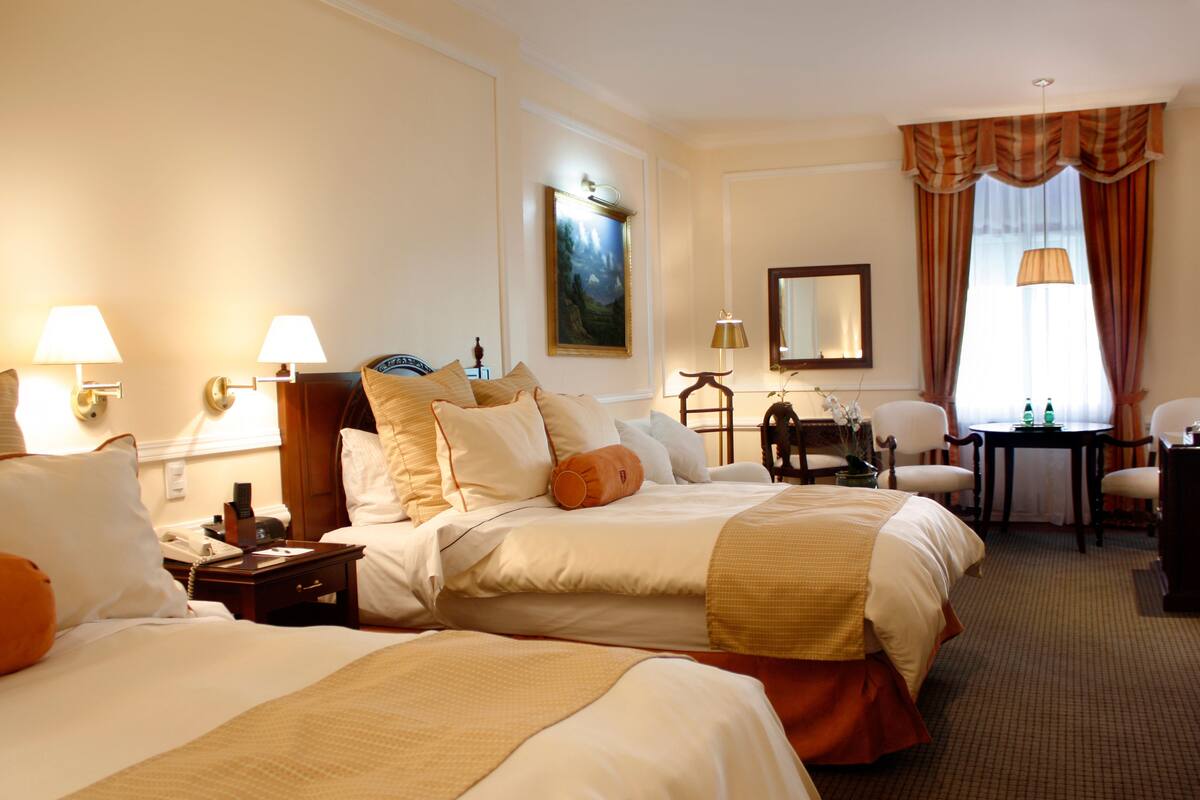 Une chambre d’hôtel avec deux lits, un bureau et une chaise. On y trouve des lampes, un téléphone et un tableau au mur.