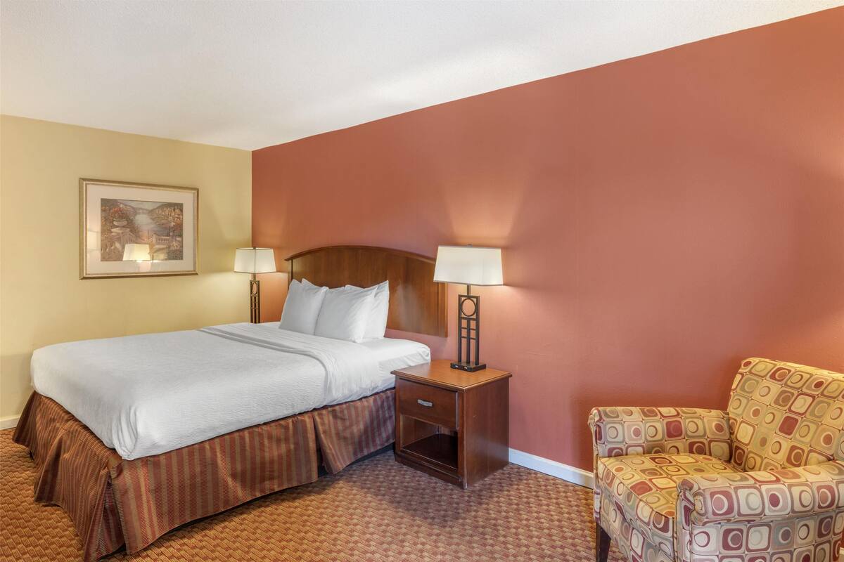 A hotel room with a bed, two bedside lamps, a chair, and a framed picture on the wall.