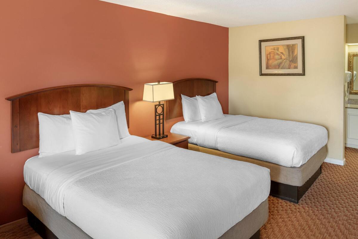 A hotel room with two beds, a nightstand with a lamp, a painting on the wall, and a bathroom visible to the right.