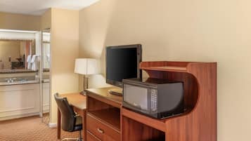 Une chambre d’hôtel équipée d’un bureau en bois, d’une télévision, d’un four à micro-ondes, d’une lampe, d’une chaise et d’une salle de bain avec un miroir et un porte-serviettes.