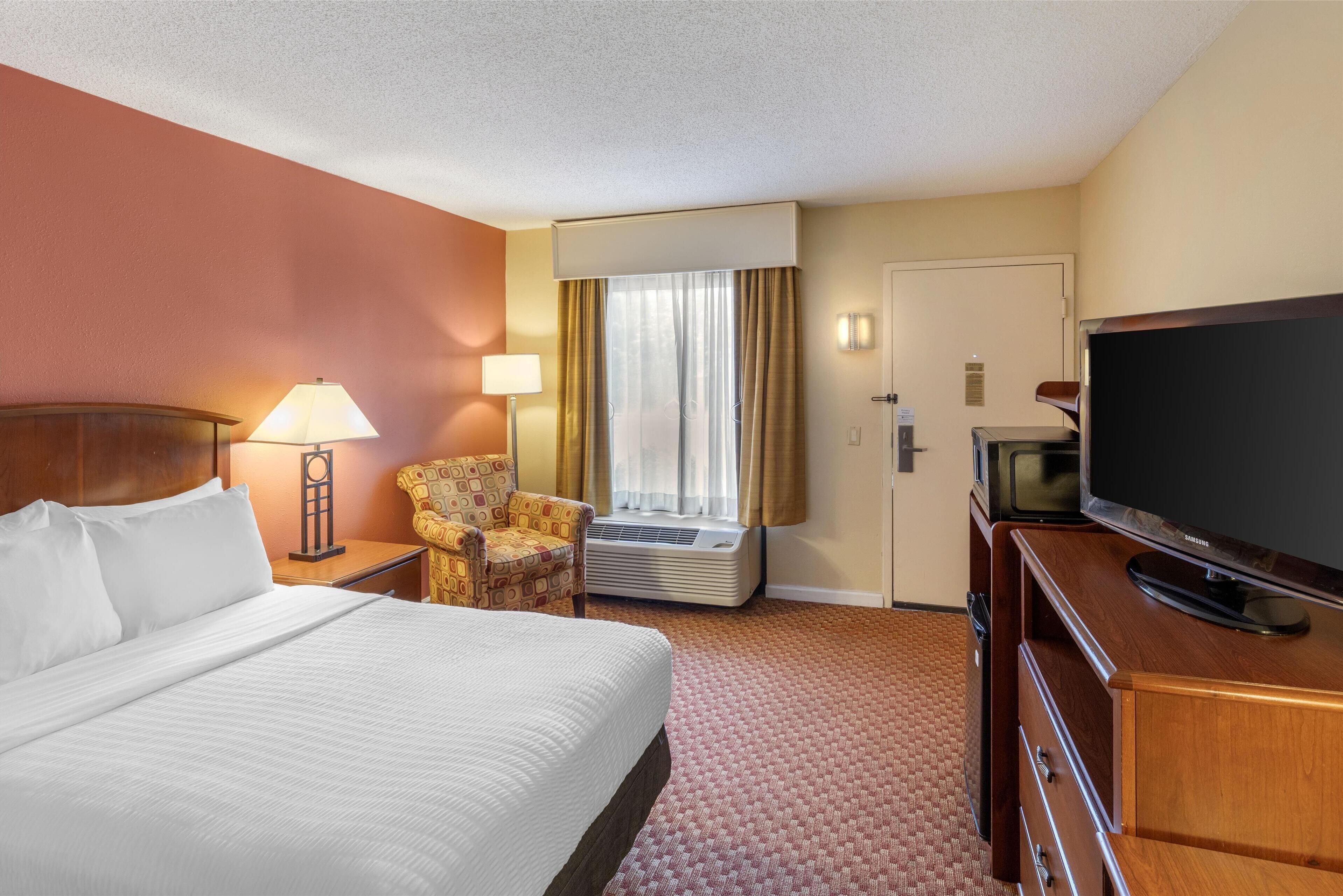 A hotel room with a large bed, a desk with a lamp, a chair, a television, and a window with curtains.