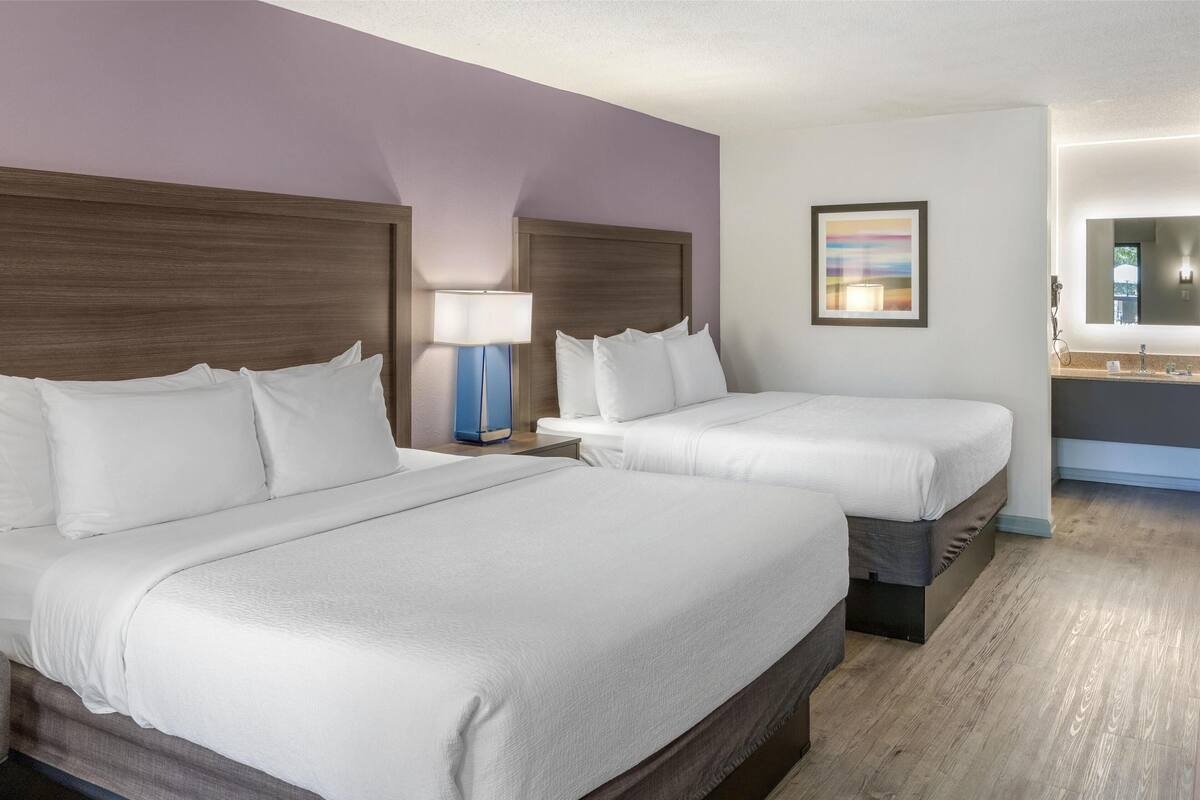 A hotel room with two beds, a nightstand with a lamp, a bathroom with a mirror, and a framed picture on the wall.