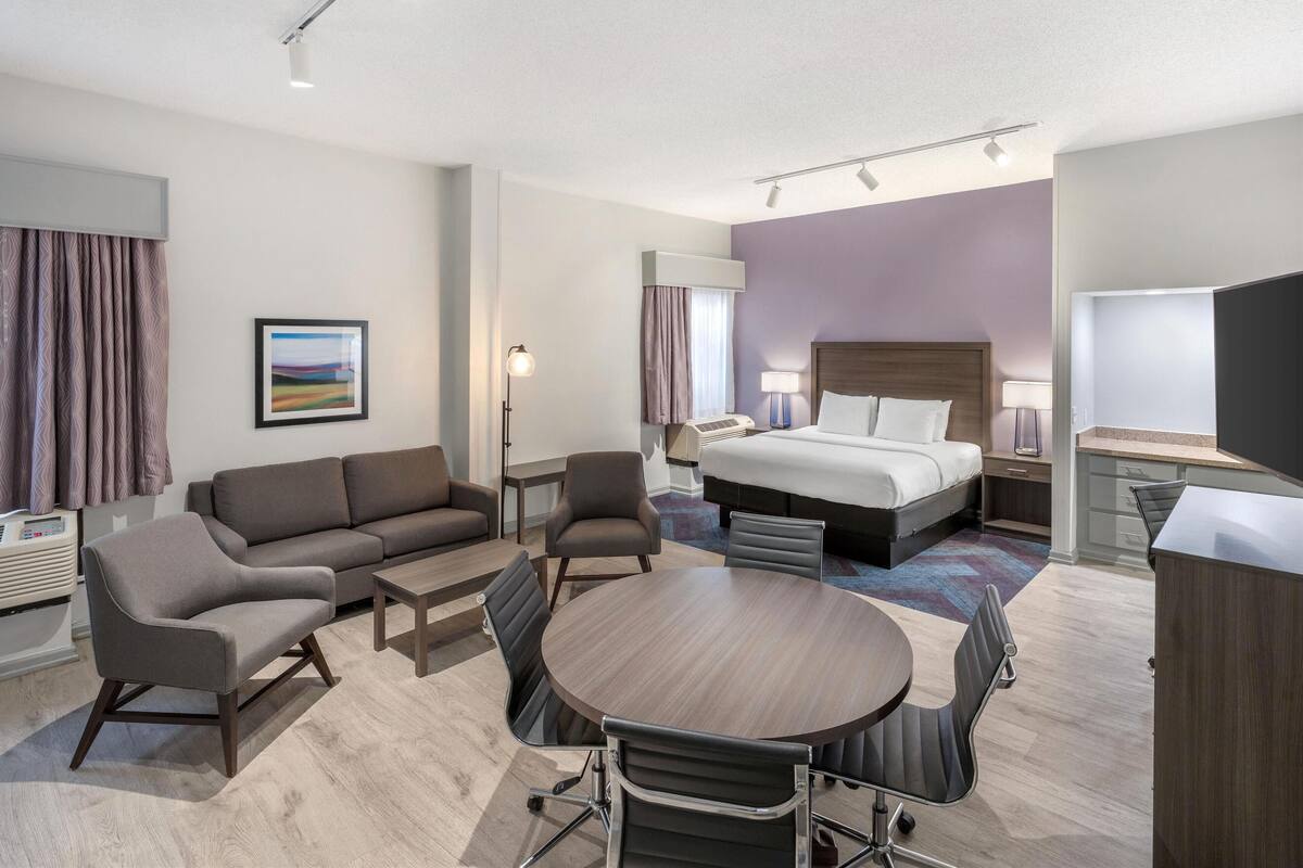 A modern hotel room with a bed, sofa, dining table, chairs, and a desk.