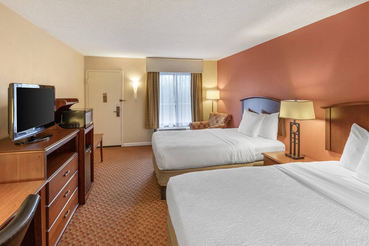 A hotel room with two beds, a desk with a television, a lamp, and a window with curtains.