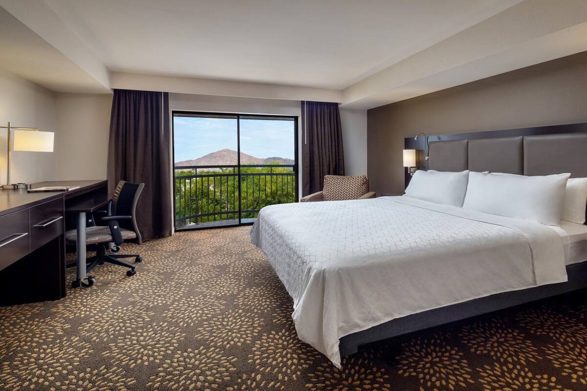 A hotel room with a large bed, a desk, a chair, and a balcony with a mountain view.