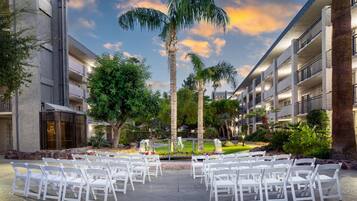 Un patio exterior con sillas blancas dispuestas para un evento, rodeado de palmeras y un edificio con balcones.