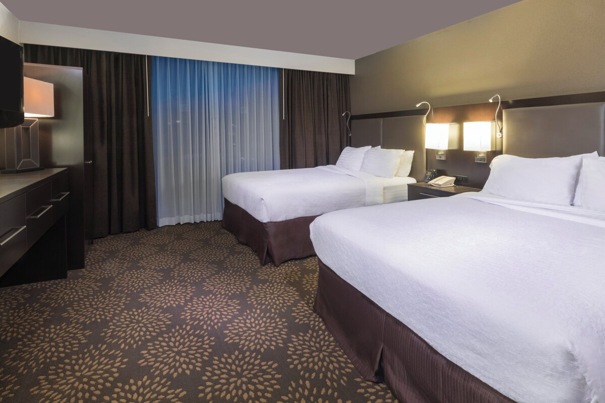 A hotel room with two beds, a desk, a TV, and a window with curtains.