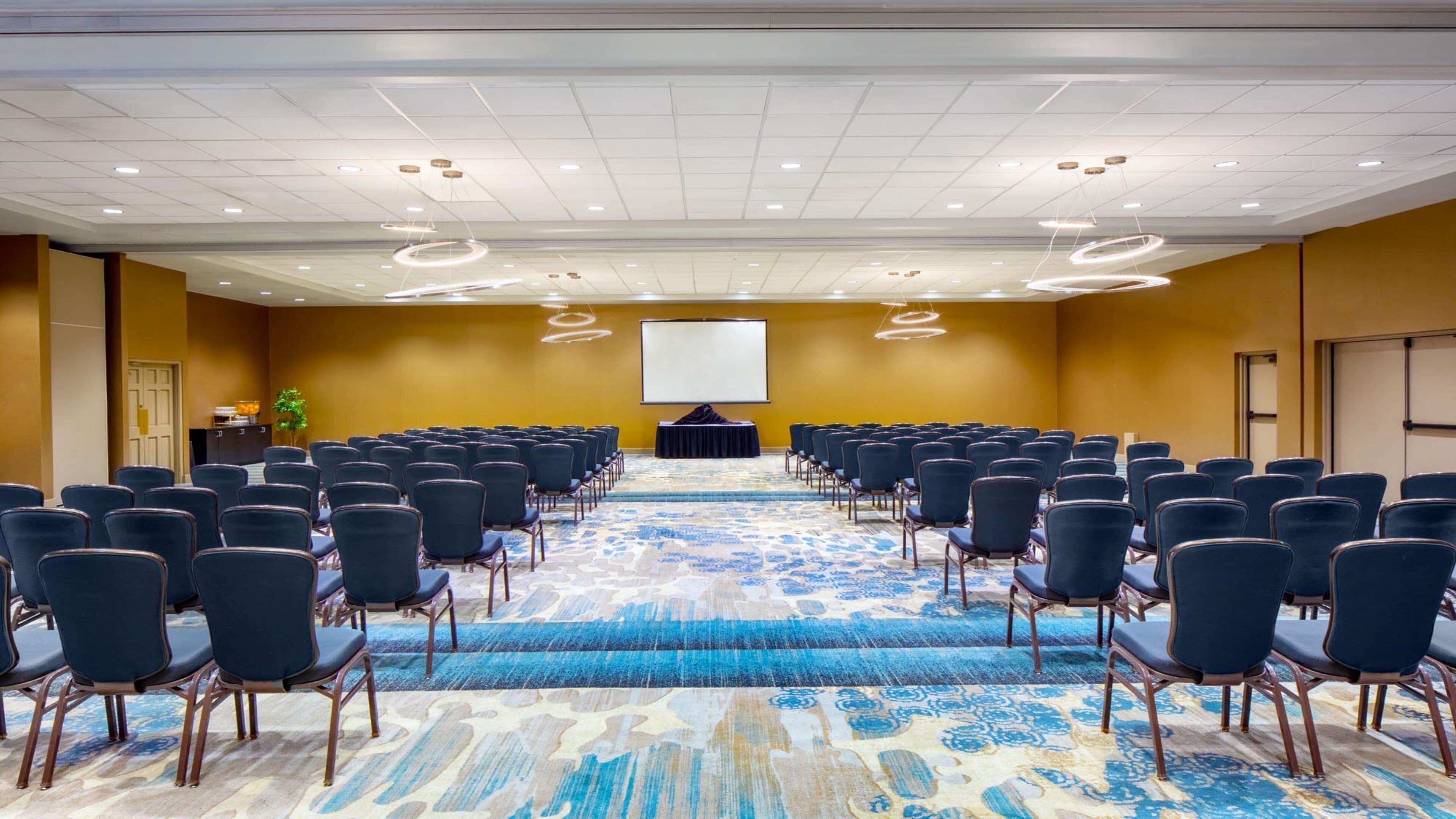 A conference room with rows of chairs, a projector screen, and a carpeted floor.