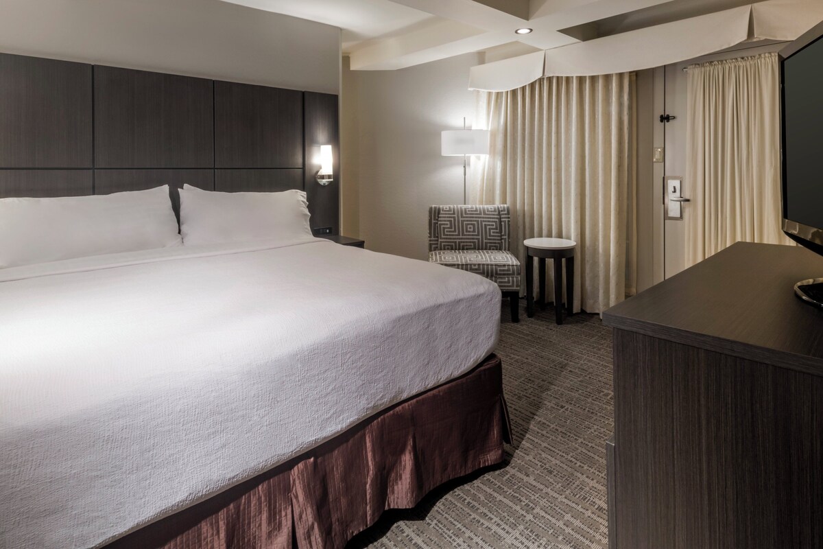 A hotel room with a large bed, a desk, a chair, a TV, and a lamp.