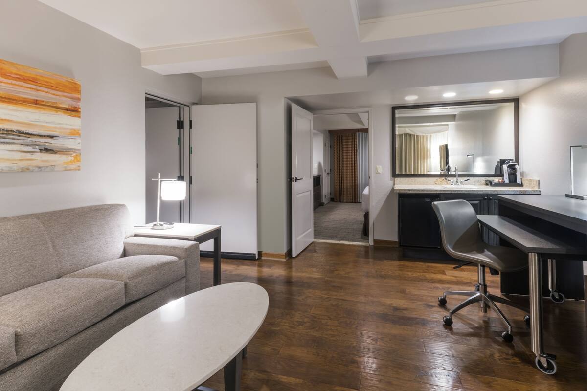 A modern hotel room with a sofa, a small table, a desk with a chair, a large mirror, and a kitchen area with a coffee machine.