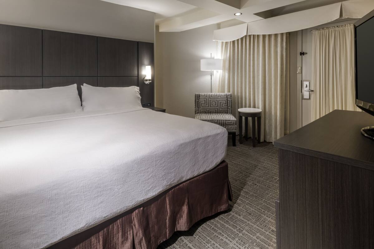 A hotel room with a large bed, a small desk, a chair, a TV, and a lamp.
