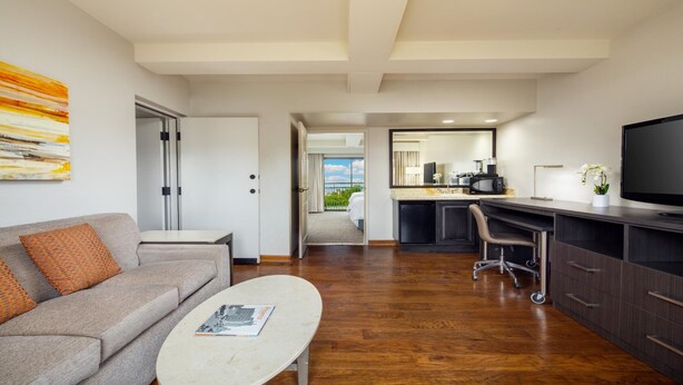 Habitación de hotel moderna con sofá, mesa de centro, televisión, kitchenette y balcón con vistas.