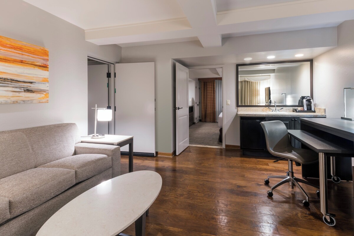 A modern hotel room with a sofa, a small table, a desk with a chair, a large mirror, and a kitchen area.