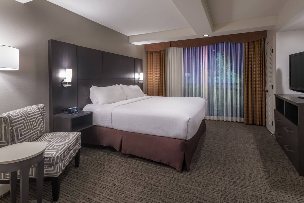 A hotel room with a large bed, a bedside table with a clock, a sofa, a TV, and a window with curtains.
