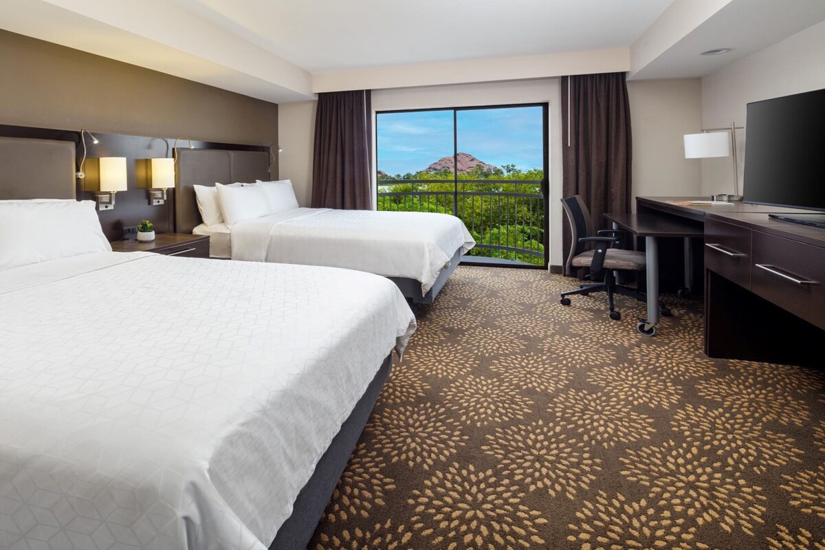 A hotel room with two beds, a desk, a chair, and a large window with a view of greenery and a mountain.