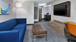 A hotel room with a blue sofa, a wooden coffee table, a TV mounted on the wall, and a small kitchenette area.