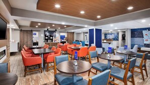 A modern hotel lobby with round tables and chairs, a bar area, and a seating area with orange and blue upholstered chairs.