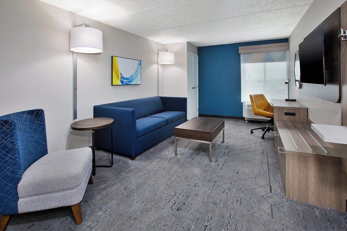 A modern hotel room with a blue sofa, a small round table, a wooden coffee table, a desk with a computer, and a TV mounted on the wall.