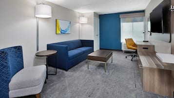 Una habitación de hotel moderna con un sofá azul, una pequeña mesa redonda, una mesa de centro de madera, un escritorio con una computadora y un televisor montado en la pared.