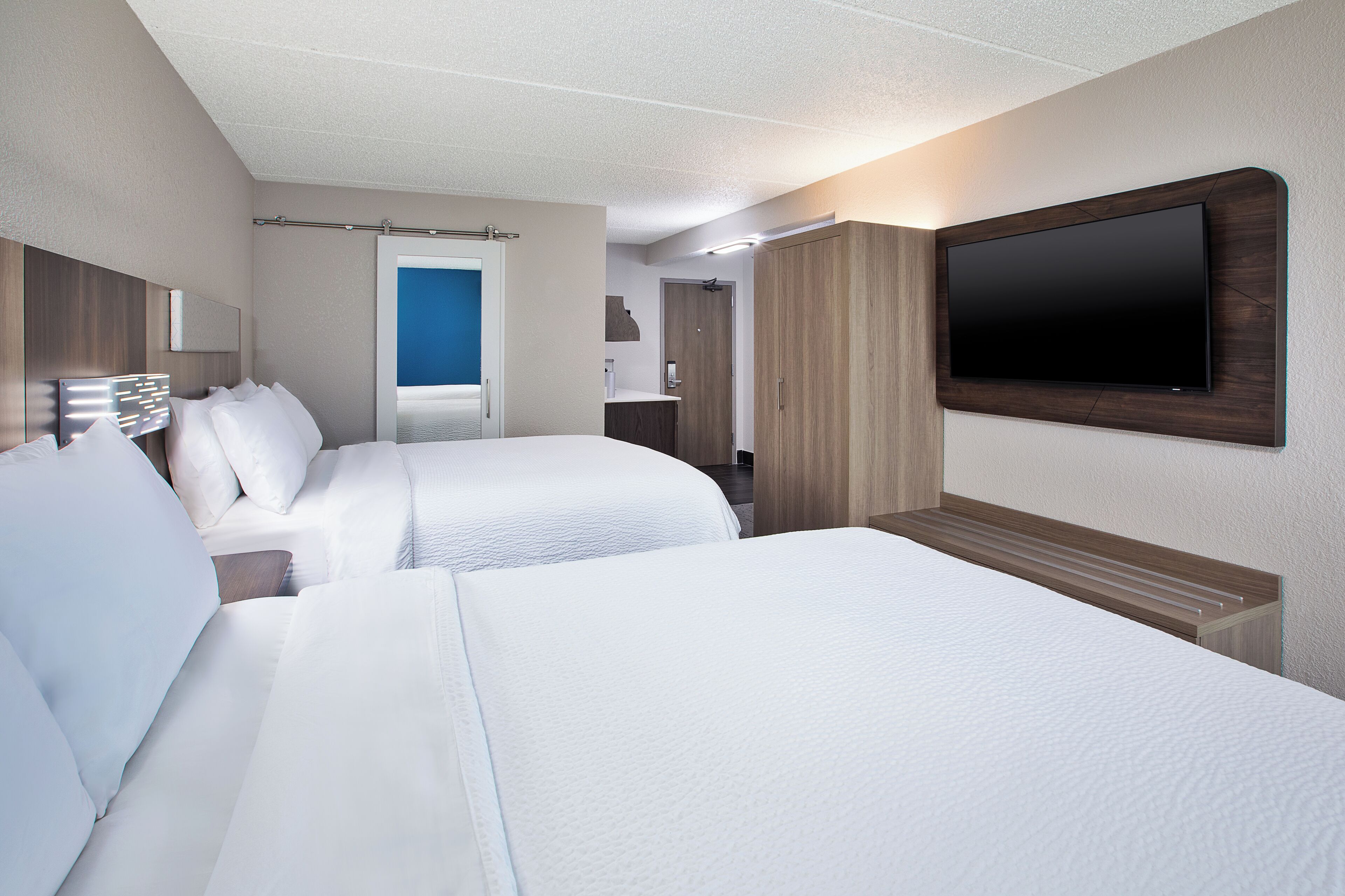 A hotel room with two beds, a flat-screen TV mounted on the wall, and a bathroom visible through an open door.