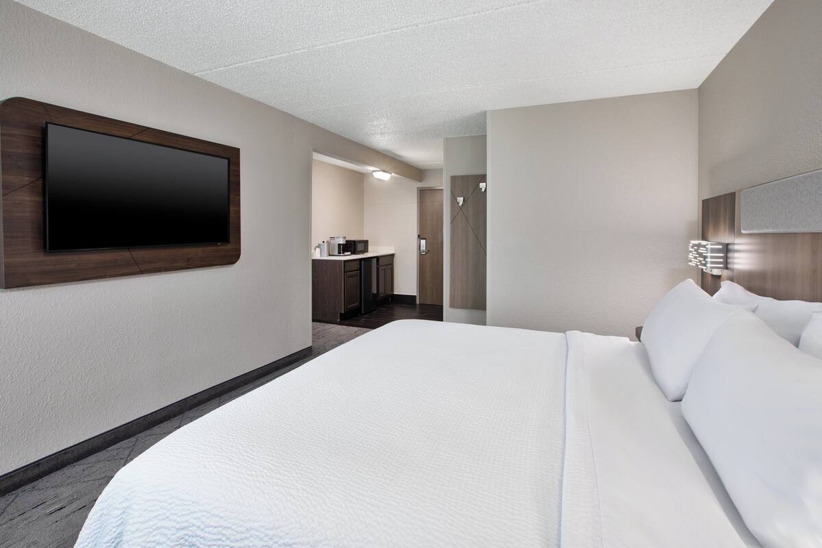 A hotel room with a large bed, a flat-screen TV mounted on the wall, a kitchenette, and a door leading to a bathroom.