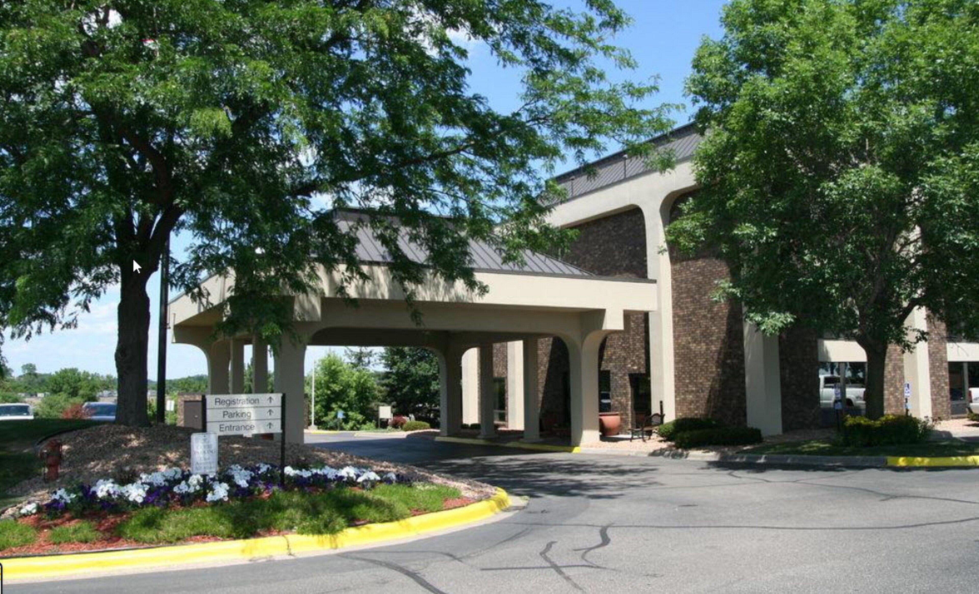 A building with a covered entrance, a sign indicating registration, parking, and entrance directions, and a landscaped area with flowers.