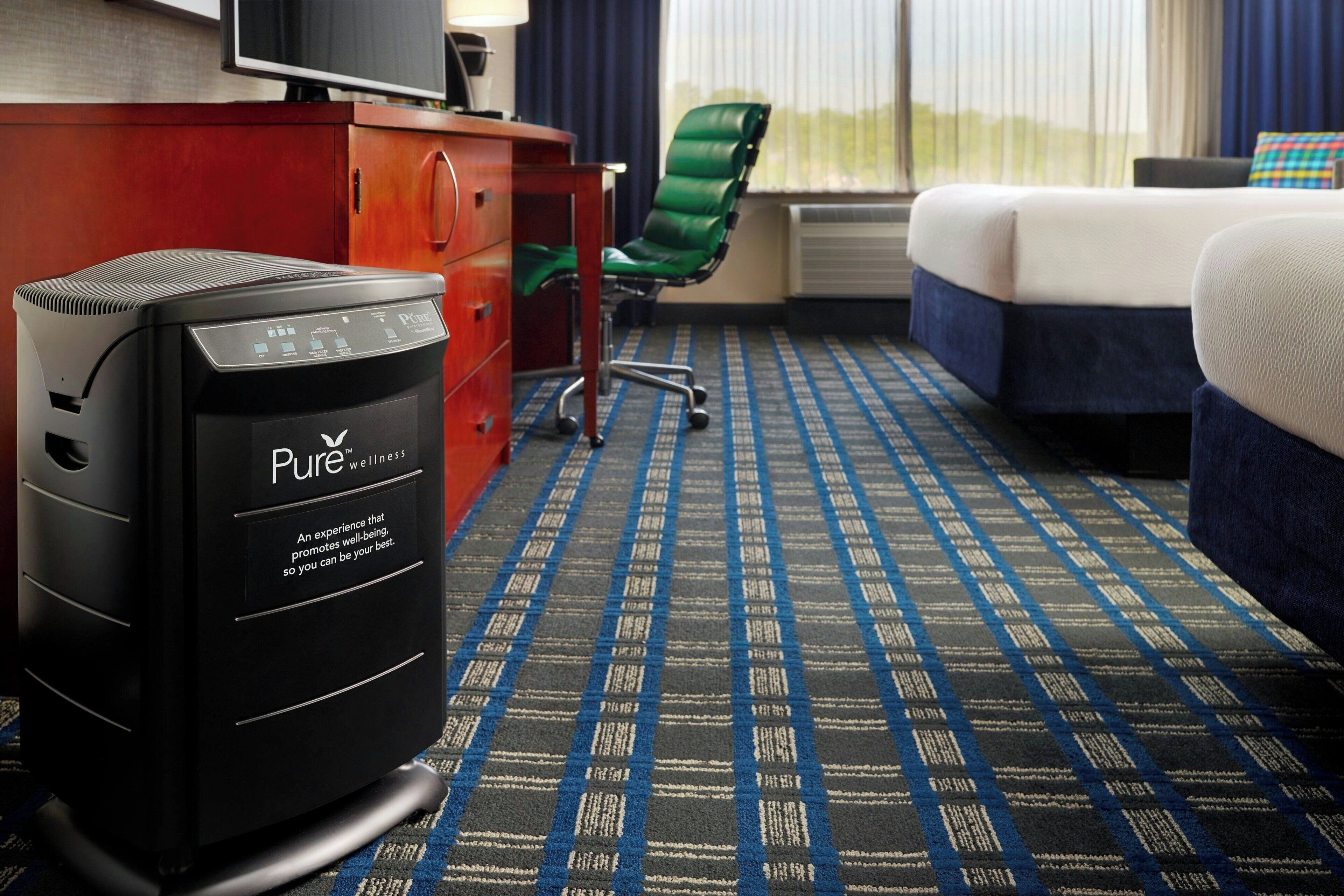 A hotel room with a bed, a desk with a computer, a green chair, and a patterned carpet.