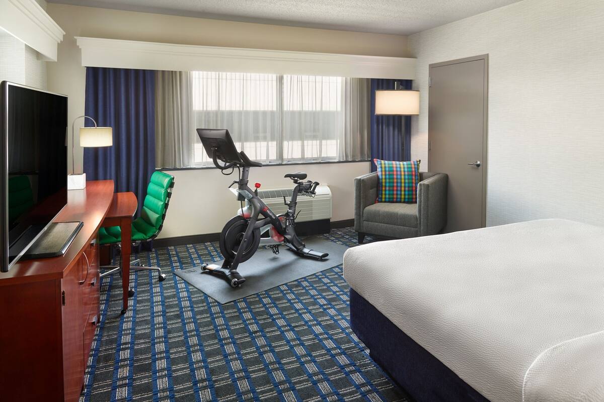 Une chambre d’hôtel comprenant un lit, un bureau avec une télévision, un vélo d’exercice, une chaise et une fenêtre avec des rideaux.